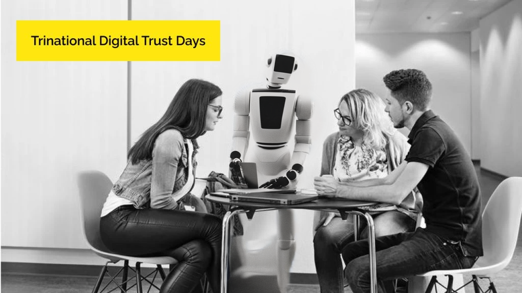 A group of three people are sitting at a table. A human-sized robot is showing a book. The people at the table are looking at what the robot is showing them..